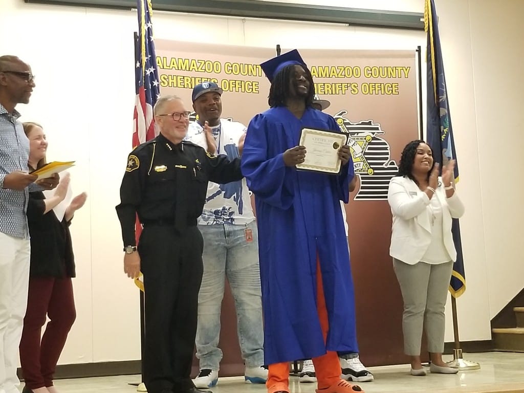 A person with orange jail jumpsuit and a blue graduation cap and gown holds a diploma while people clap.